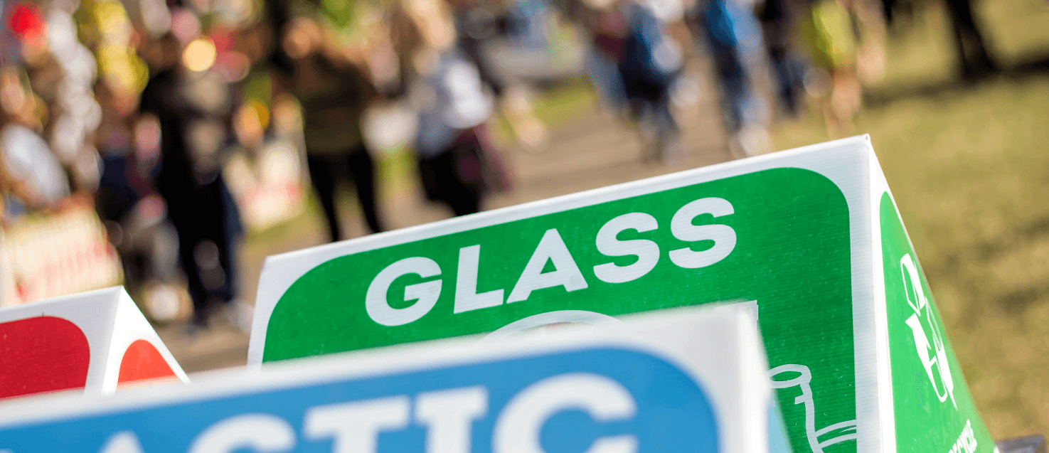A focused close up photo of a green glass recycling bin and blue plastic ahead of it, blurred int he background are people walking on a field
